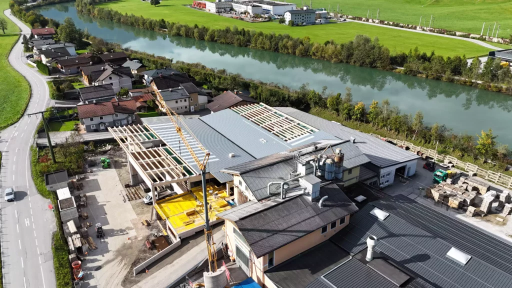 default Luftaufnahme einer Baustelle mit Hallenbau und Kran an einem Flussufer neben Wohnhäusern und grünen Wiesen