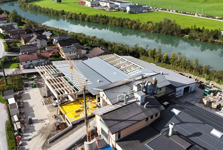 Luftaufnahme einer Baustelle mit Hallenbau und Kran an einem Flussufer neben Wohnhäusern und grünen Wiesen