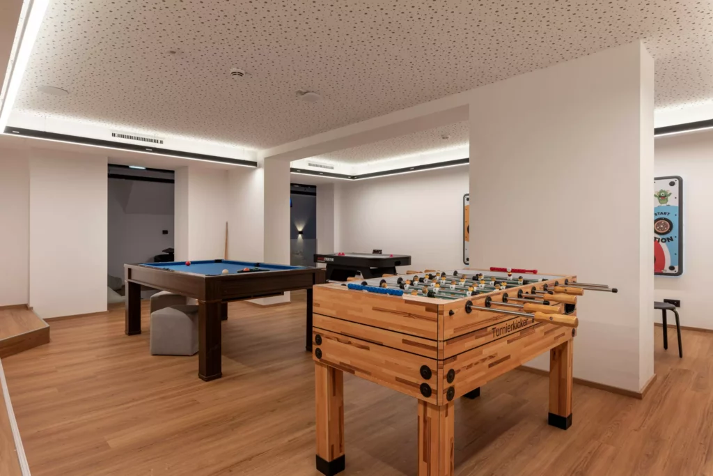 2024-06-13_Deutinger-Hotel-Kesselgrub-©sobietzki-high-res-13 Moderner Spielraum mit Kicker, Billardtisch und Airhockey in einem Hotel mit Holzfußboden und weißer Decke