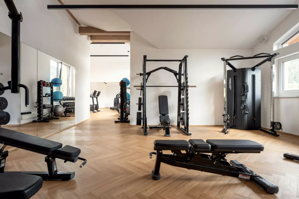 2024-06-13_Deutinger-Hotel-Kesselgrub-©sobietzki-high-res-15 Moderner Fitnessraum mit Kraftstationen, Hantelbänken und Spiegelwand in hellem Hotelinnenraum mit Holzfußboden