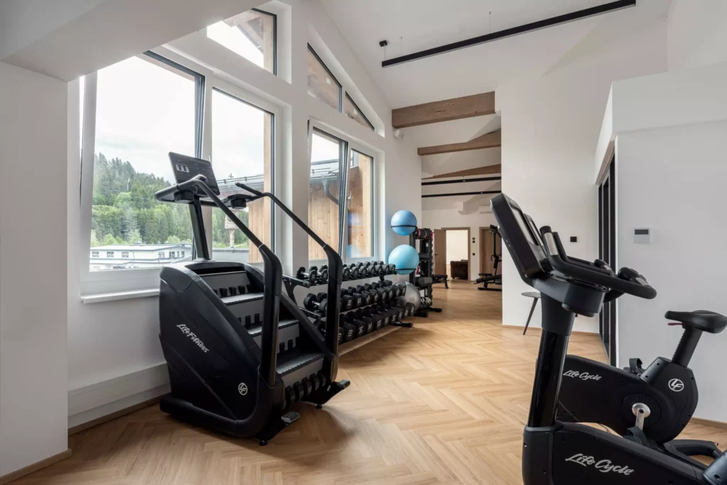 2024-06-13_Deutinger-Hotel-Kesselgrub-©sobietzki-high-res-16 Moderner Fitnessraum mit Crosstrainer, Heimtrainer, Hanteln und Gymnastikbällen in hellem Raum mit großen Fenstern und