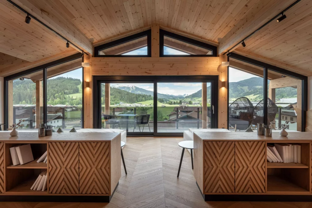 2024-06-13_Deutinger-Hotel-Kesselgrub-©sobietzki-high-res-18 Modernes Hotelzimmer mit Holzdecke, zwei Tischen, Stühlen und großem Panoramafenster mit Bergblick im Deutinger Hotel