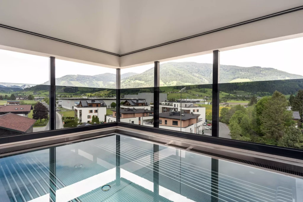 2024-06-13_Deutinger-Hotel-Kesselgrub-©sobietzki-high-res-25 Überdachter Außenpool mit Glasgeländer und Blick auf moderne Wohnhäuser und grüne Berge im Hintergrund