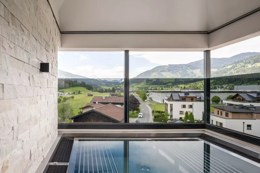 2024-06-13_Deutinger-Hotel-Kesselgrub-©sobietzki-high-res-26 Blick von einem modernen Balkon mit Glasgeländer und Whirlpool auf ein Tal mit Häusern und bewaldeten Bergen im Hintergrund
