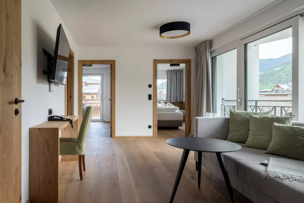 2024-06-13_Deutinger-Hotel-Kesselgrub-©sobietzki-high-res-32 Modern eingerichtetes Hotelzimmer mit Sofa, grünem Stuhl, Flachbildfernseher und Blick auf Berglandschaft durch große Fenster