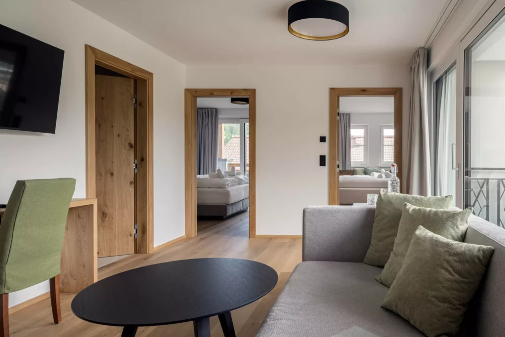 2024-06-13_Deutinger-Hotel-Kesselgrub-©sobietzki-high-res-33 Modern eingerichtetes Hotelzimmer mit Sofa, grünem Sessel, Holztüren und Blick auf zwei Schlafzimmer im Deutinger Hotel