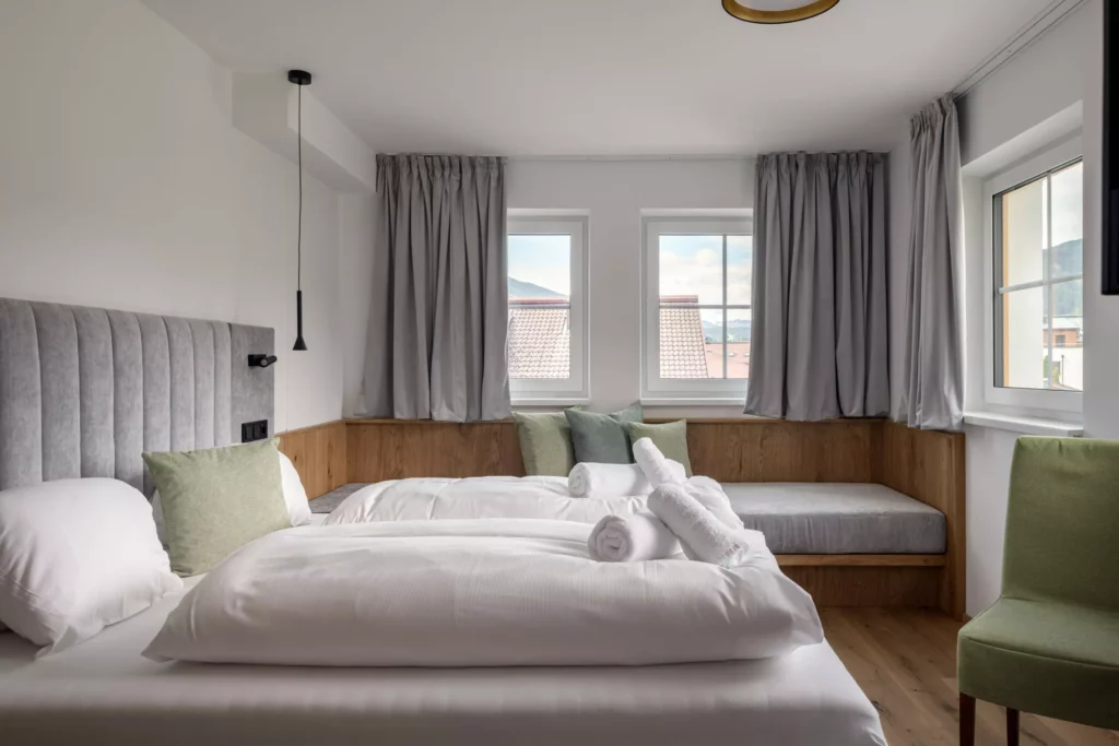 2024-06-13_Deutinger-Hotel-Kesselgrub-©sobietzki-high-res-34 Modern eingerichtetes Hotelzimmer mit Doppelbett, grauem Kopfteil, grünen Kissen und Fensterbank mit Sitzkissen und Ausblick