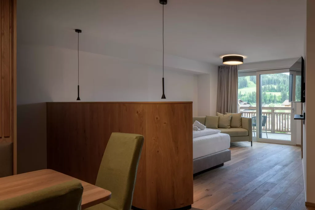 2024-06-13_Deutinger-Hotel-Kesselgrub-©sobietzki-high-res-36 Modern eingerichtetes Hotelzimmer mit Bett, Sofa, Holzmöbeln und Balkon mit Bergblick