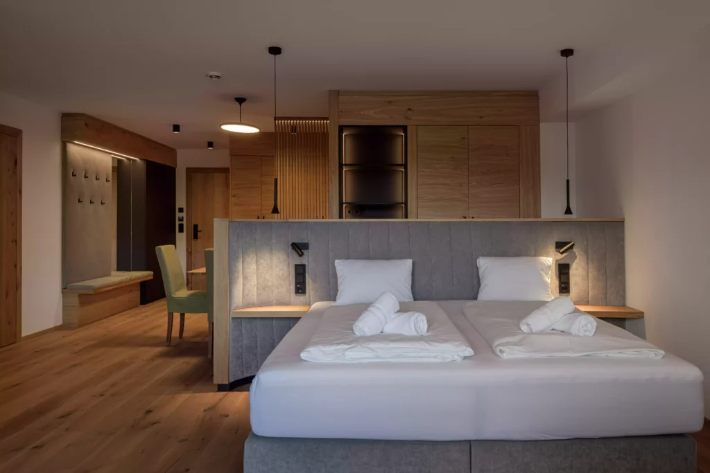 2024-06-13_Deutinger-Hotel-Kesselgrub-©sobietzki-high-res-37 Modern eingerichtetes Hotelzimmer mit Doppelbett, Holzboden, Sitzbereich und integrierter Küchenzeile im Hintergrund