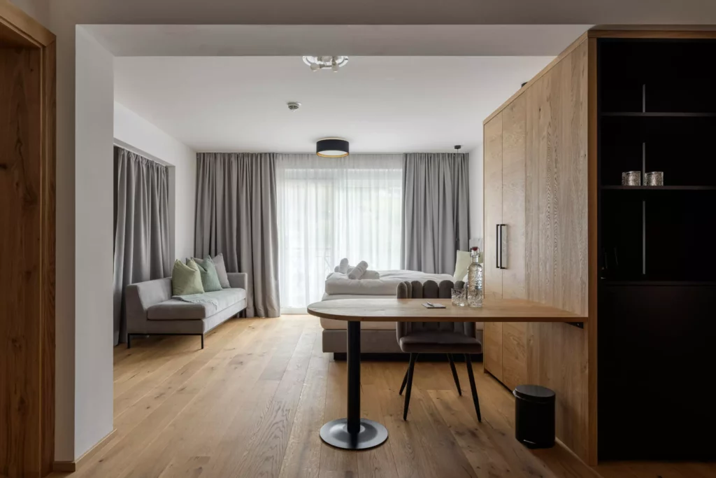2024-06-13_Deutinger-Hotel-Kesselgrub-©sobietzki-high-res-38 Modern eingerichtetes Hotelzimmer mit Holzboden, Sofa, Bett, Tisch und großem Fenster mit Vorhängen im Deutinger Hotel
