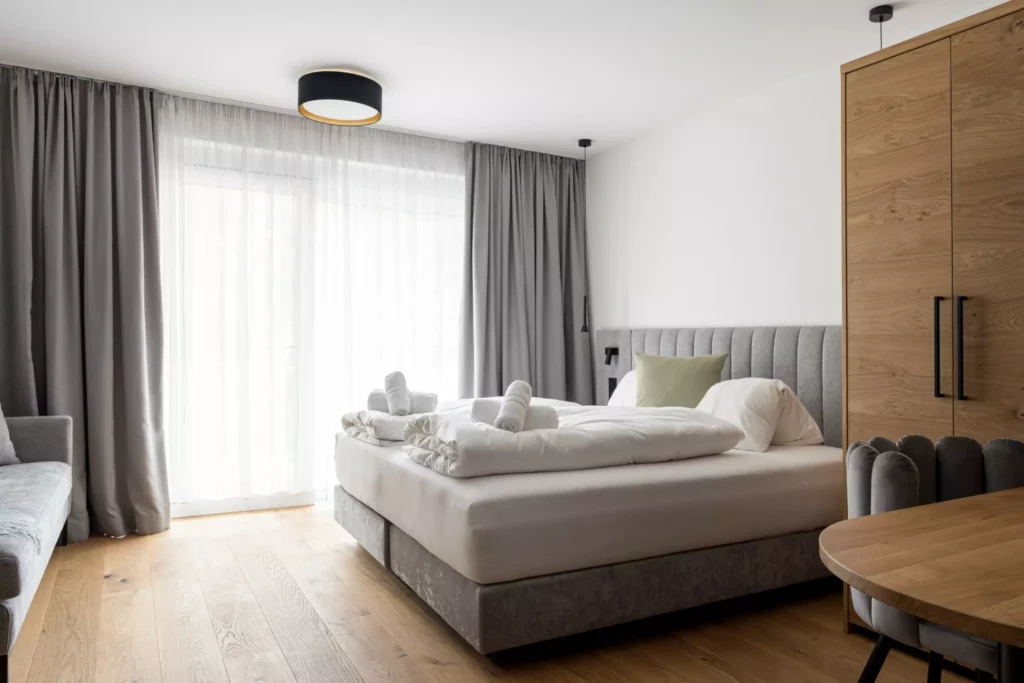 2024-06-13_Deutinger-Hotel-Kesselgrub-©sobietzki-high-res-39 Modern eingerichtetes Hotelzimmer mit Doppelbett, grauem Kopfteil, Holzschrank, Sofa und großem Fenster mit Vorhängen