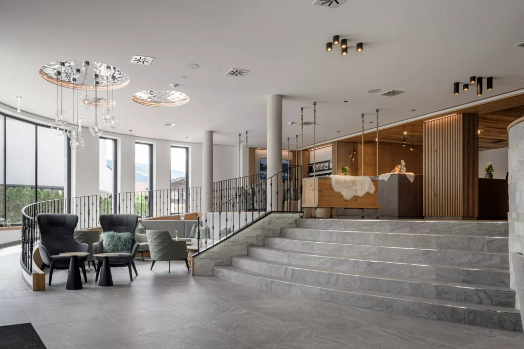 2024-06-13_Deutinger-Hotel-Kesselgrub-©sobietzki-high-res-40 Moderne Hotellobby mit Sitzgruppen, großen Fenstern, Treppe und Holz-Elementen im Deutinger Hotel Kesselgrub Innenbereich.