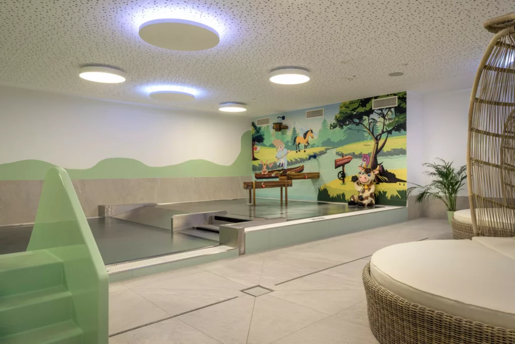 2024-06-13_Deutinger-Hotel-Kesselgrub-©sobietzki-high-res-9 Innenraum eines Kinderhallenbads mit Rutsche, Wandmalerei mit Tieren und gemütlichen Sitzgelegenheiten im Hotel Kesselgrub