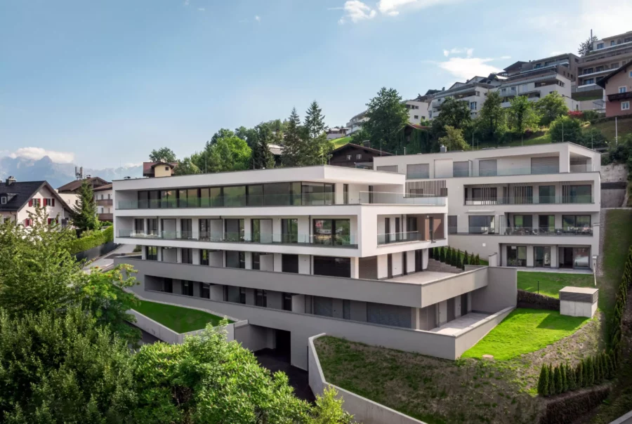 Moderne Wohnanlage mit klarer Architektur auf Hanglage, umgeben von Grünflächen und Nachbarhäusern, bei klarem Himmel.