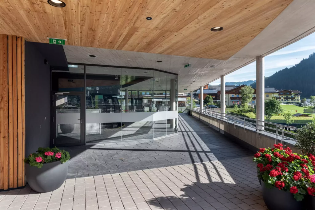 2024-08-19_Talstation-Kieserl-Grossarl-©sobietzki-low-res-2560px-17 Moderne Talstation mit Glasfront, Holzdecke und Blumendekoration in Großarl, umgeben von Bergen und Gebäuden im Hintergrund.