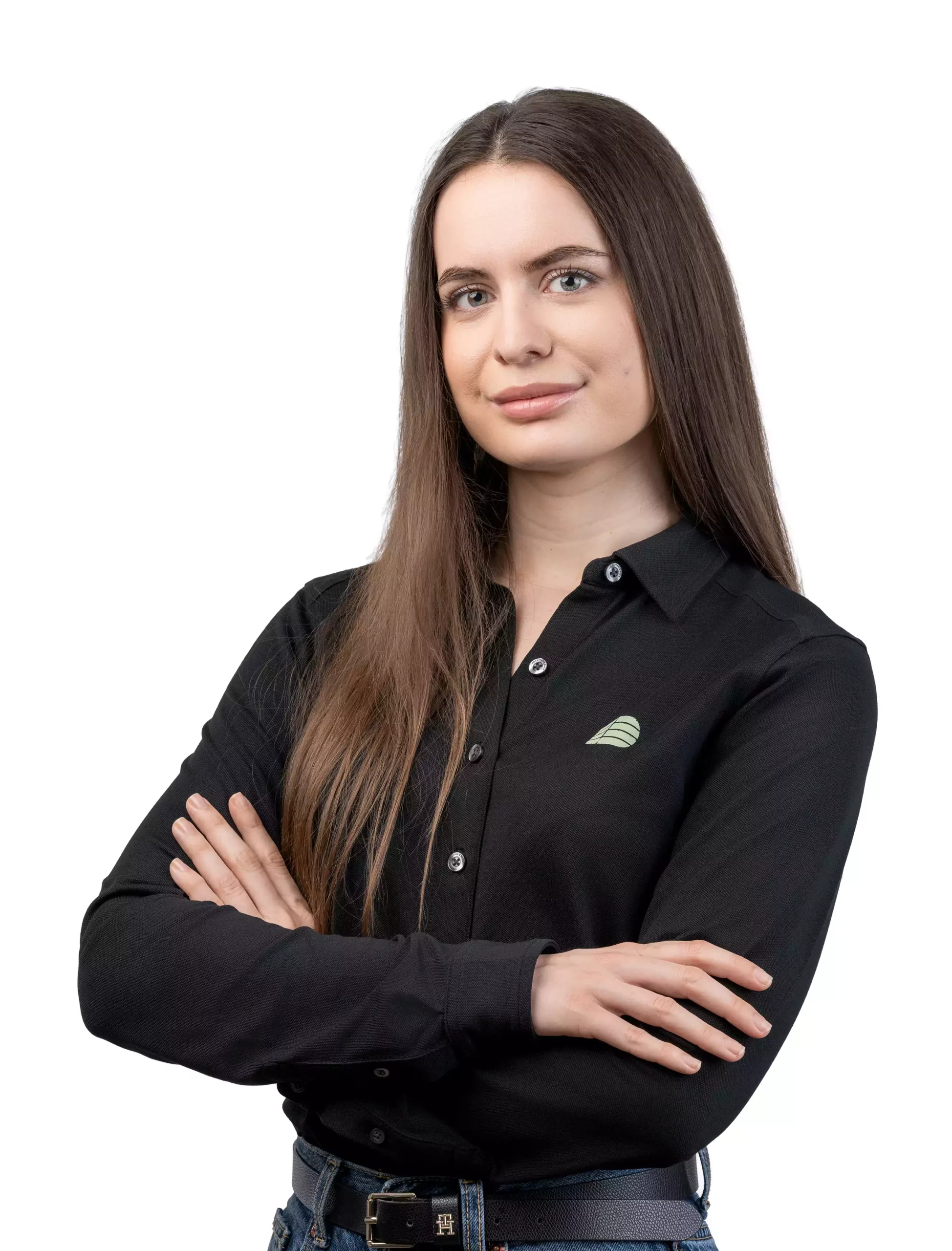 Junge Frau mit langen braunen Haaren trägt schwarze Bluse mit Logo, Arme verschränkt, vor weißem Hintergrund.
