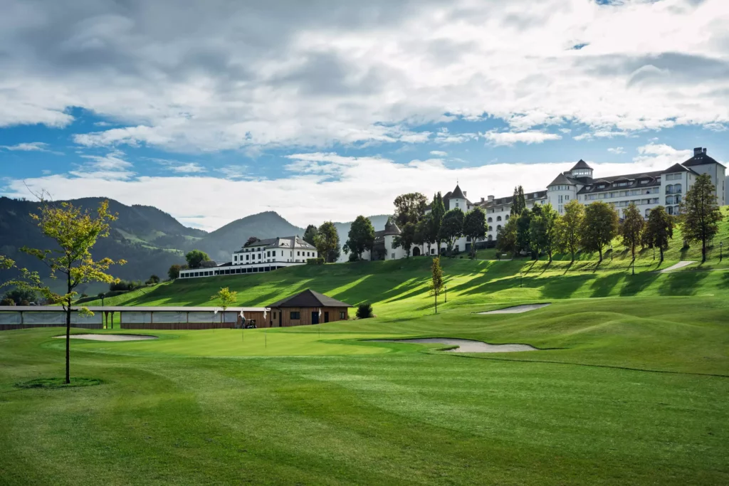 251016_Deutinger-Mitarbeiterhaus-Pichlarn-©sobietzki-low-res-2560px-1 Grünes Golfplatzgelände mit Bunker, Bäumen und einem großen weißen Gebäude im Hintergrund unter bewölktem Himmel in Pichlarn