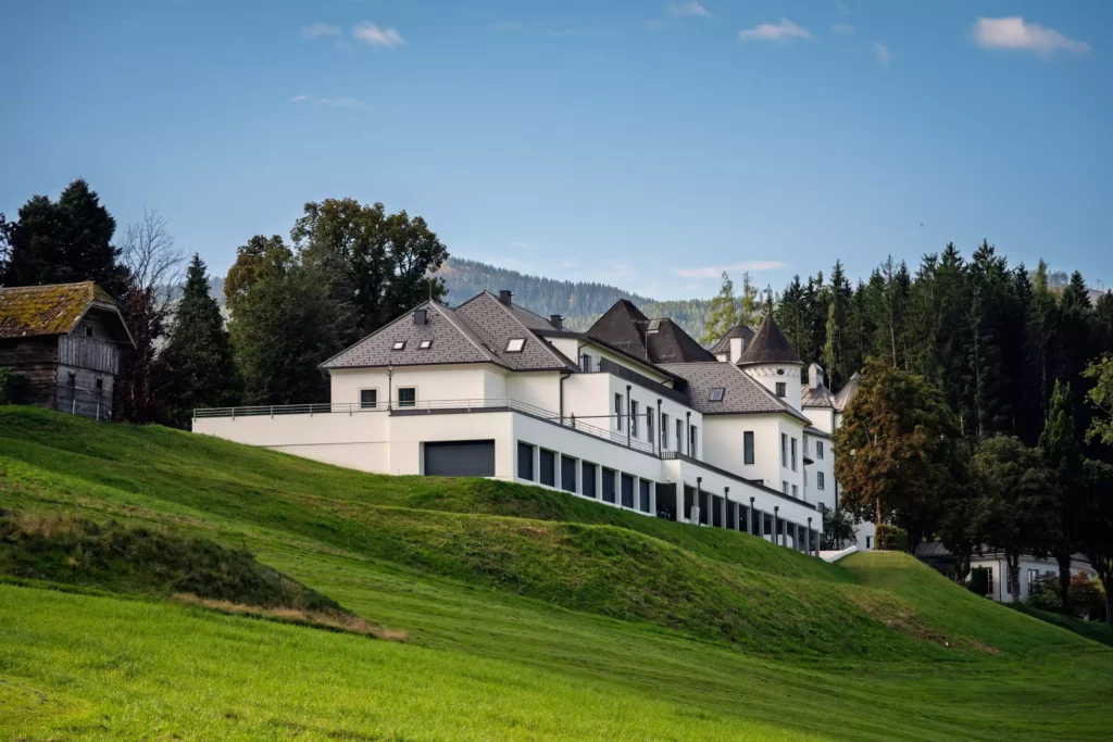 251016_Deutinger-Mitarbeiterhaus-Pichlarn-©sobietzki-low-res-2560px-10 Modernes weißes Mitarbeiterhaus auf grünem Hügel mit Wald und Bergen im Hintergrund bei klarem Himmel