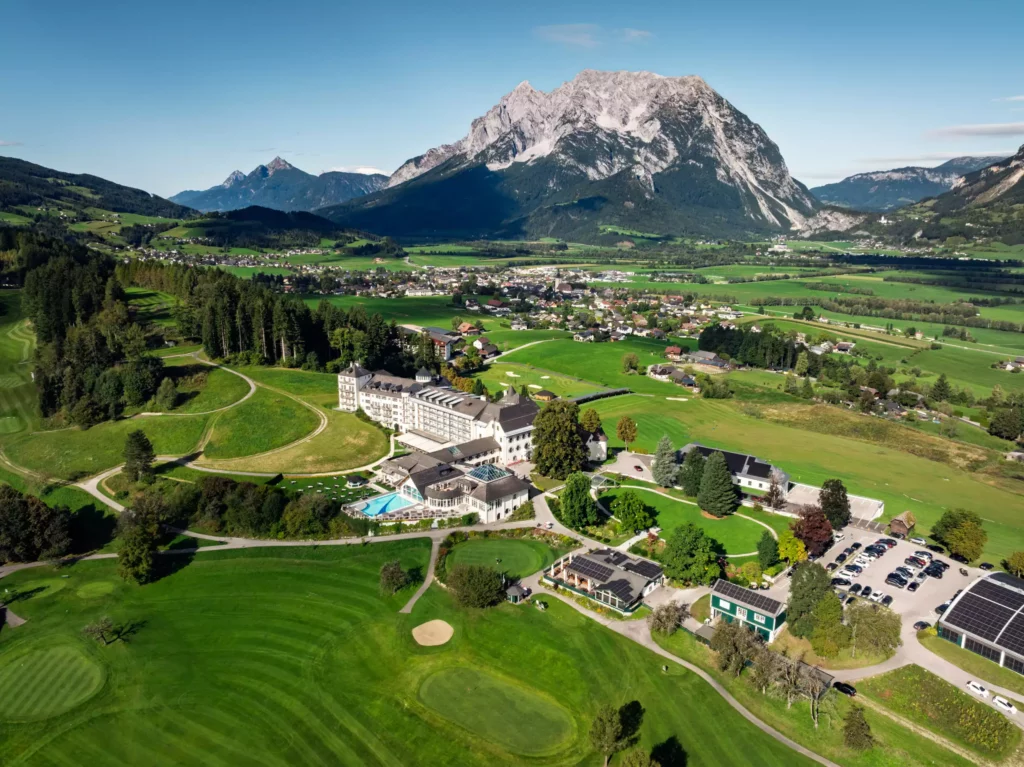 251016_Deutinger-Mitarbeiterhaus-Pichlarn-©sobietzki-low-res-2560px-18 Luftaufnahme eines großen Hotelkomplexes mit Pool, umgeben von Golfplatz, Wiesen und Bergen im Hintergrund bei klarem Himmel