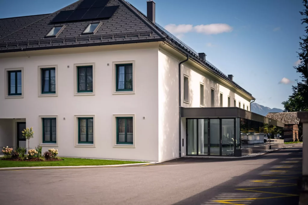 251016_Deutinger-Mitarbeiterhaus-Pichlarn-©sobietzki-low-res-2560px-8 Modernes zweistöckiges Mitarbeiterhaus mit weißer Fassade, grünen Fenstern und Glasanbau bei sonnigem Himmel in Pichlarn