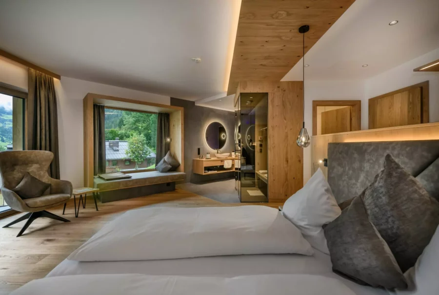 Modern eingerichtetes Hotelzimmer mit großem Bett, gemütlichem Sessel, Fensterbank mit Sitzkissen und Blick auf grüne