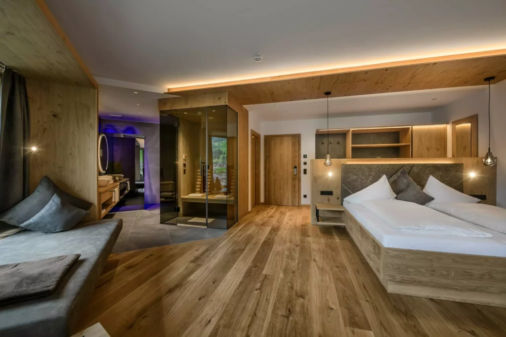 Hubertushof_03 Modern eingerichtetes Hotelzimmer Hubertushof mit Holzfußboden, Doppelbett, Sitzbank und Glas-Sauna im Hintergrund