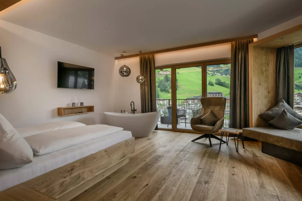 Hubertushof_04 Modernes Hotelzimmer mit Doppelbett, freistehender Badewanne, Sessel, Balkon und Blick auf grüne Hügel im Hubertushof