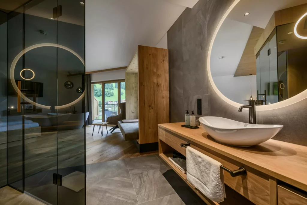 Hubertushof_05 Modernes Badezimmer mit beleuchtetem Rundspiegel, Holzwaschtisch und Blick in einen Wohnbereich mit Balkon und Berglandschaft