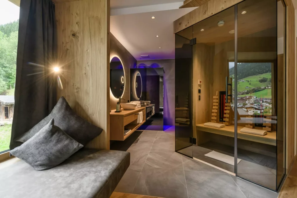 Hubertushof_06 Modernes Badezimmer mit Sauna, großem Fenster mit Bergblick, Sitzbank mit Kissen und beleuchtetem Spiegel im Hubertushof