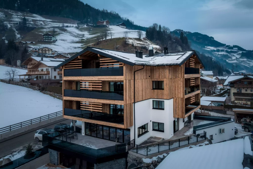 deutinger projektentwicklung baumanagement generalunternehmen immobilien grossarl hallein salzburg Diellehen-©sobietzki-high-res-4 Modernes Holz- und Weißbauwerk mit großen Balkonen vor schneebedeckten Bergen und Häusern in Großarl, Salzburg