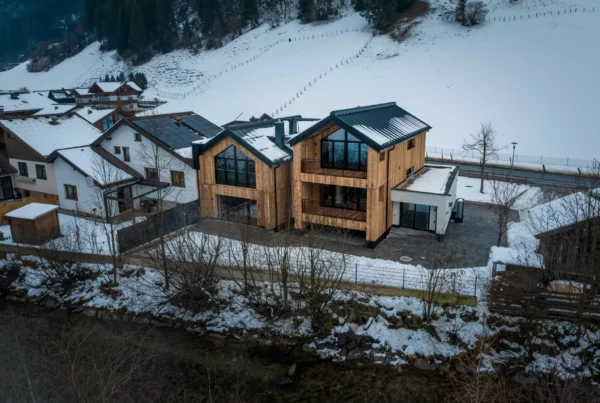 Zwei moderne Holzhäuser mit großen Fenstern und Balkonen in verschneiter Berglandschaft neben einem Bachlauf und weiteren
