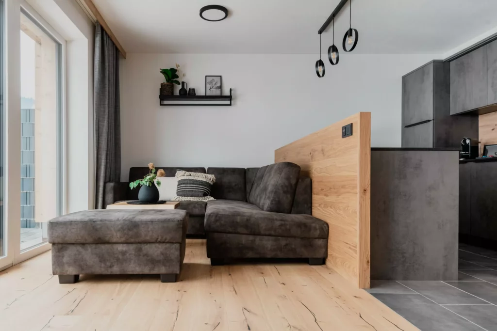deutinger projektentwicklung baumanagement generalunternehmen immobilien grossarl hallein salzburg Hettegger-Sandra-©sobietzki-high-res-7 Modern eingerichtetes Wohnzimmer mit grauer Couch, Hocker, Holzboden und offener Küche mit grauen Fronten und Holztrennwand