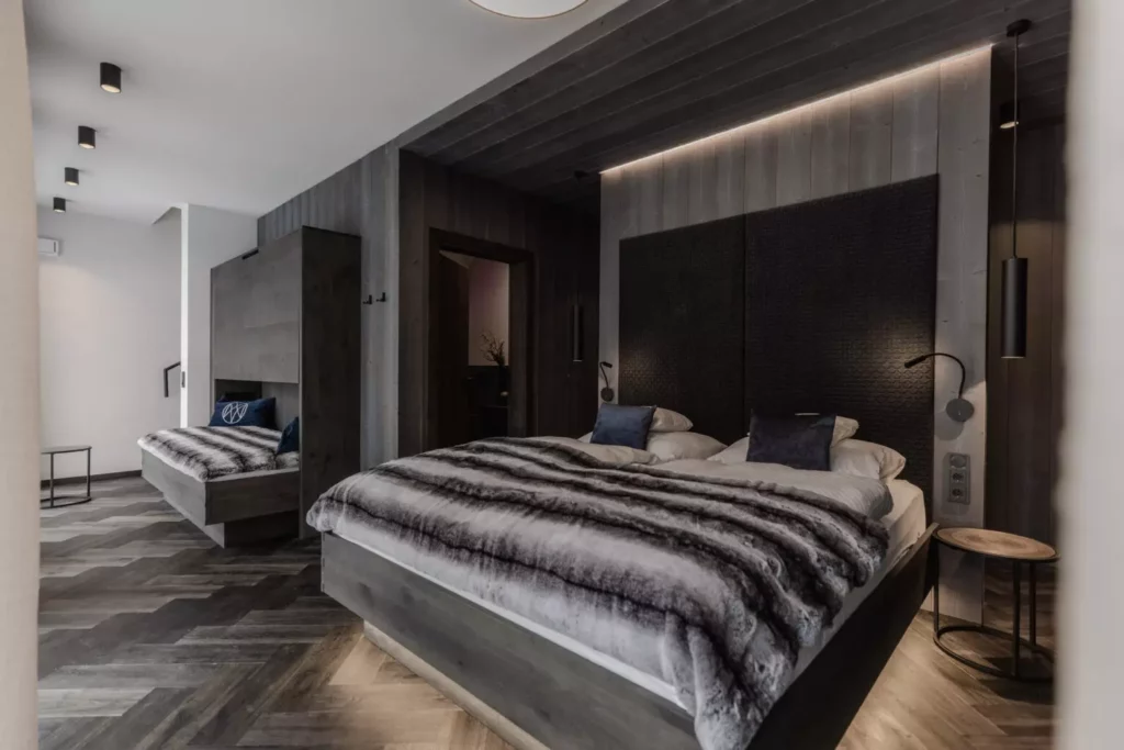 deutinger-projektentwicklung-baumanagement-generalunternehmen-immobilien-grossarl-hallein-salzburg-_77A5377-© Foto by studio manufaktur2 Modern eingerichtetes Schlafzimmer mit Doppelbett, grauem Fellüberwurf, dunklen Holzwänden und indirekter Beleuchtung