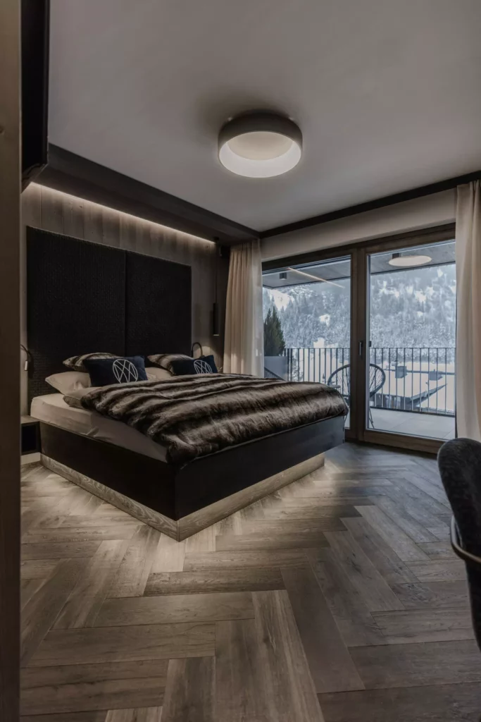 deutinger-projektentwicklung-baumanagement-generalunternehmen-immobilien-grossarl-hallein-salzburg-_77A5394-© Foto by studio manufaktur2 Modern eingerichtetes Schlafzimmer mit großem Bett, Holzboden, bodentiefen Fenstern und Blick auf verschneite Berge