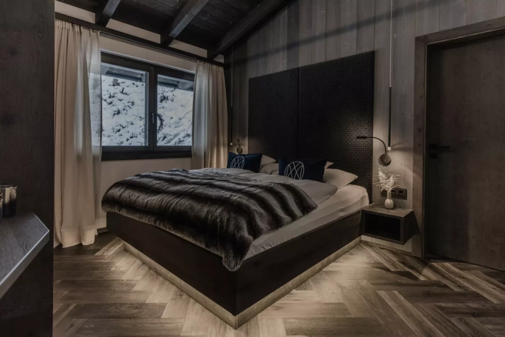 deutinger-projektentwicklung-baumanagement-generalunternehmen-immobilien-grossarl-hallein-salzburg-onkl-xonna-155-© Foto by studio manufaktur2 Modernes Schlafzimmer mit großem Bett, dunklem Holz, Fellüberwurf und Blick auf verschneite Landschaft durch Fenster