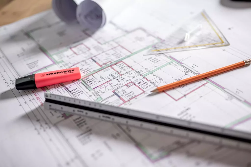 deutinger-projektentwicklung-baumanagement-generalunternehmen-immobilien-salzburg-68 Architekturplan mit orangem Bleistift, rosa Stabilo Boss Textmarker, Dreiecklineal und Maßstab auf Holztisch