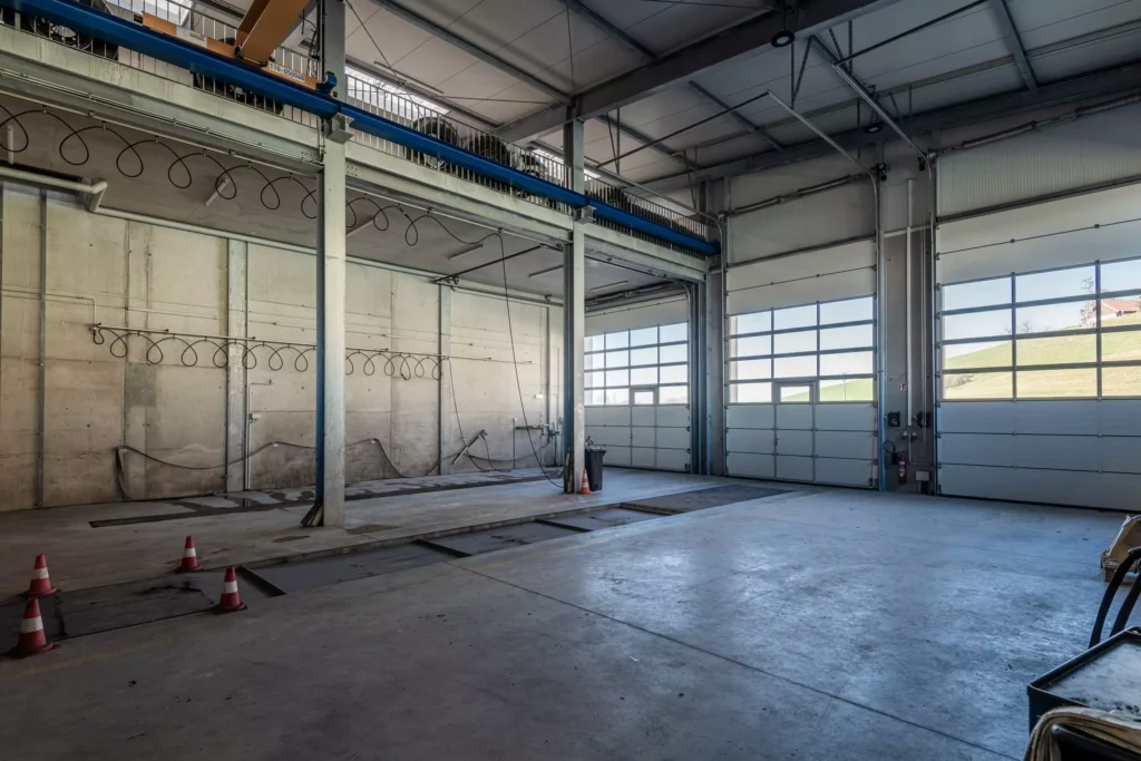 deutinger-projektentwicklung-baumanagement-generalunternehmen-immobilien-salzburg-Betriebshalle-Meister-Hallwang-11 Leere Betriebshalle mit großen Garagentoren und Betonboden in Salzburg, geeignet für Baumanagement und Bauprojektentwicklung.
