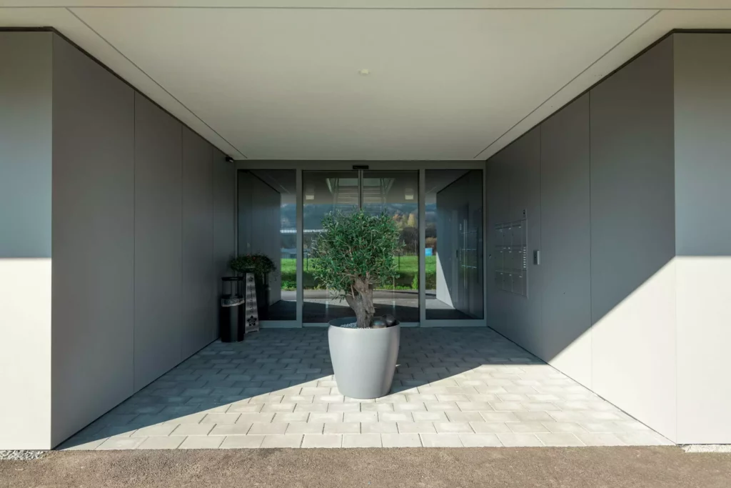 deutinger-projektentwicklung-baumanagement-generalunternehmen-immobilien-salzburg-Betriebshalle-Meister-Hallwang-14 Moderner Eingangsbereich mit grauen Wänden, Pflanze in großem Topf und Glastür bei Betriebshalle Meister Hallwang