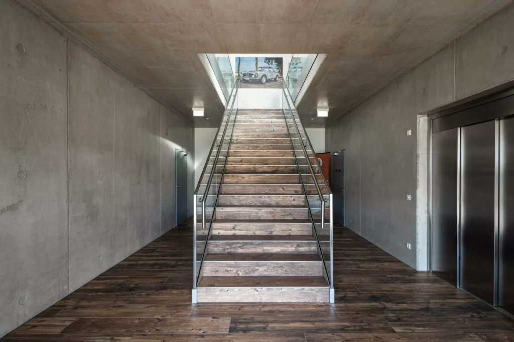 deutinger-projektentwicklung-baumanagement-generalunternehmen-immobilien-salzburg-Betriebshalle-Meister-Hallwang-15 Moderner Innenraum mit Holztreppe, Glasgeländer, Betonwänden und Aufzug in einer Betriebshalle in Hallwang bei Salzburg