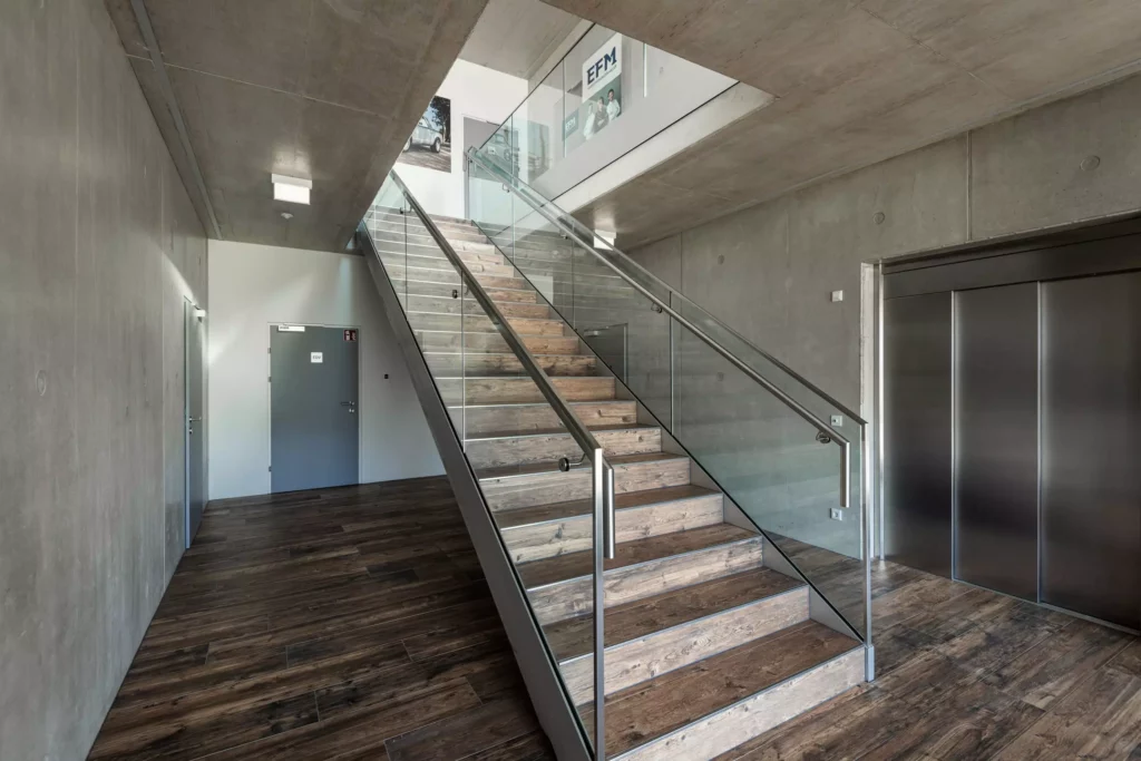 deutinger-projektentwicklung-baumanagement-generalunternehmen-immobilien-salzburg-Betriebshalle-Meister-Hallwang-16 Moderner Innenraum mit Holztreppe, Glasgeländer, Betonwänden und Aufzug in einer Betriebshalle in Hallwang bei Salzburg