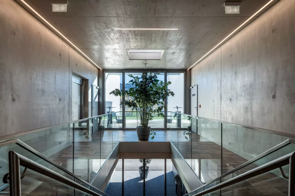 deutinger-projektentwicklung-baumanagement-generalunternehmen-immobilien-salzburg-Betriebshalle-Meister-Hallwang-17 Moderner Innenraum mit Treppe, Glasgeländer, Betonwänden und großen Fenstern in einer Betriebshalle in Hallwang bei Salzburg