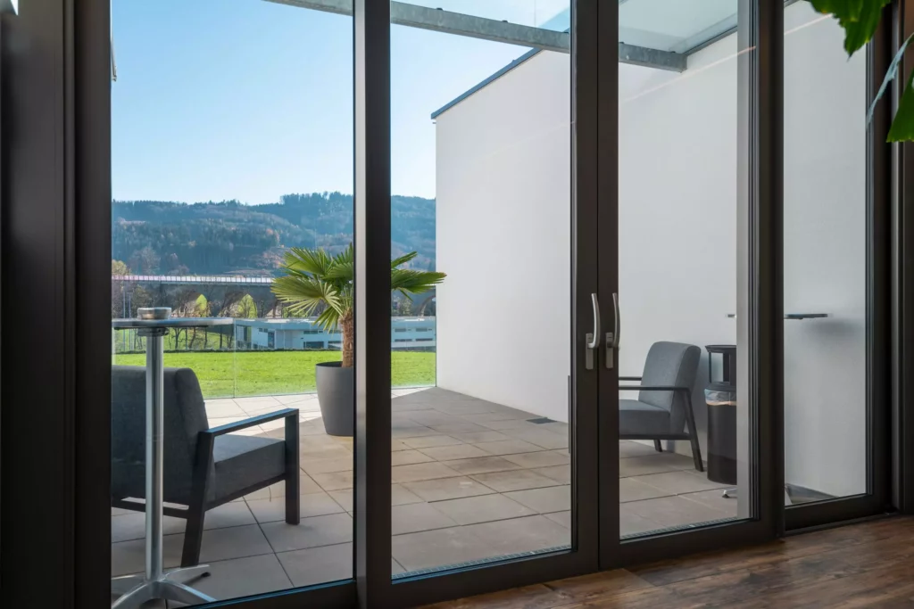 deutinger-projektentwicklung-baumanagement-generalunternehmen-immobilien-salzburg-Betriebshalle-Meister-Hallwang-18 Blick durch große Glas-Schiebetüren auf Terrasse mit zwei grauen Sesseln, Pflanze und grüner Wiese vor bewaldeten Hügeln