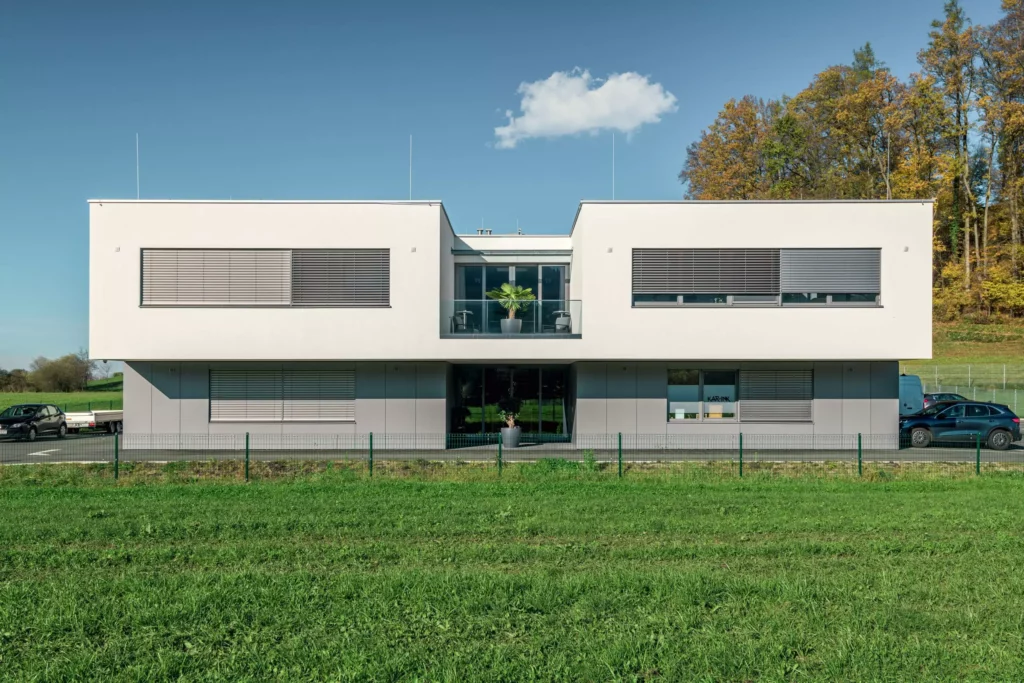 deutinger-projektentwicklung-baumanagement-generalunternehmen-immobilien-salzburg-Betriebshalle-Meister-Hallwang-2 Moderne zweigeschossige Betriebshalle mit großen Fenstern, Balkon und Parkplätzen vor grünem Feld bei klarem Himmel