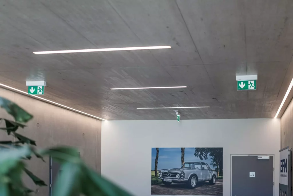 deutinger-projektentwicklung-baumanagement-generalunternehmen-immobilien-salzburg-Betriebshalle-Meister-Hallwang-20 Moderner Innenraum mit Betonwänden und Decke, Notausgangsschildern und Bild eines klassischen Mercedes Cabriolets an der Wand