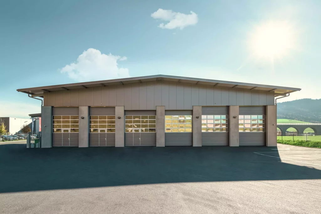 deutinger-projektentwicklung-baumanagement-generalunternehmen-immobilien-salzburg-Betriebshalle-Meister-Hallwang-3 Moderne Betriebshalle mit sechs großen Sektionaltoren unter klarem Himmel bei Sonnenschein in Hallwang nahe Salzburg