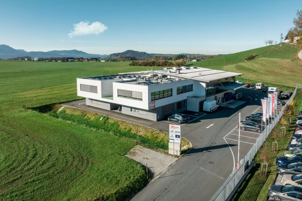 deutinger-projektentwicklung-baumanagement-generalunternehmen-immobilien-salzburg-Betriebshalle-Meister-Hallwang-4 Moderne Betriebshalle mit Parkplätzen und Firmenflaggen in ländlicher Umgebung bei Hallwang, Salzburg, unter klarem Himmel