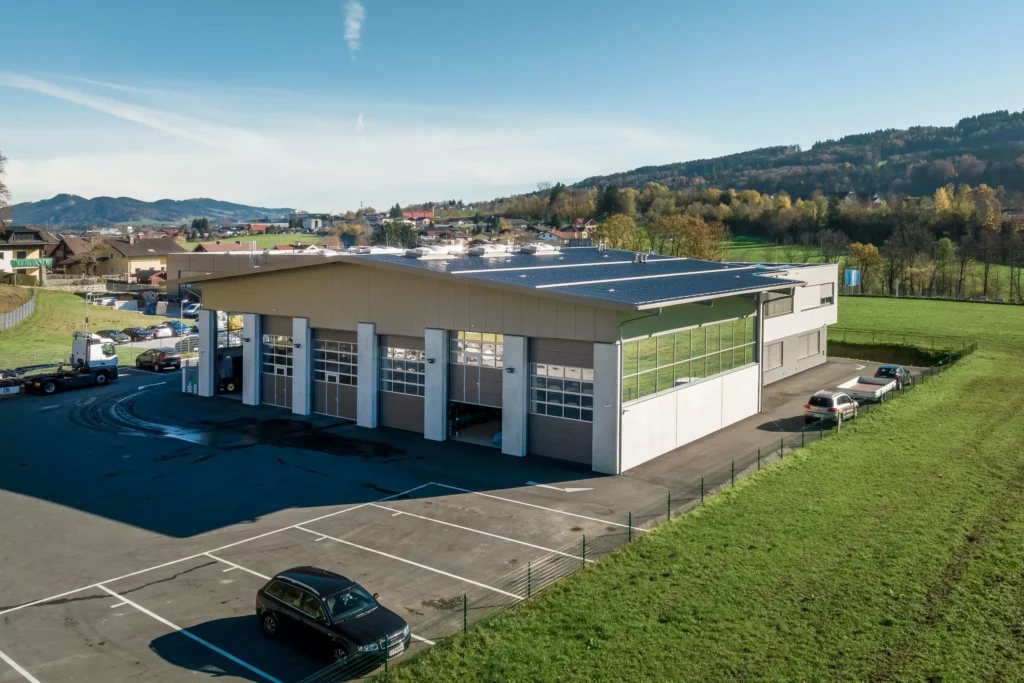 deutinger-projektentwicklung-baumanagement-generalunternehmen-immobilien-salzburg-Betriebshalle-Meister-Hallwang-5 Moderne Betriebshalle mit Solarpaneelen auf dem Dach, umgeben von Parkplätzen und grüner Landschaft in Hallwang bei Salzburg.
