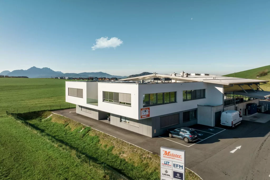 deutinger-projektentwicklung-baumanagement-generalunternehmen-immobilien-salzburg-Betriebshalle-Meister-Hallwang-6 Moderne Betriebshalle mit Parkplatz und Firmenbeschilderung vor grüner Wiese und Bergen bei klarem Himmel in Hallwang