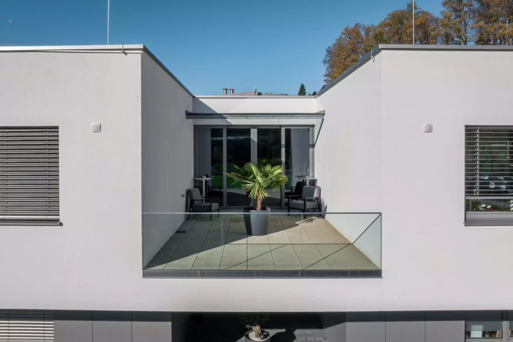 deutinger-projektentwicklung-baumanagement-generalunternehmen-immobilien-salzburg-Betriebshalle-Meister-Hallwang-7 Moderner Balkon mit Glasgeländer, zwei schwarzen Sesseln und einer großen Topfpflanze an einem weißen Gebäude bei klarem