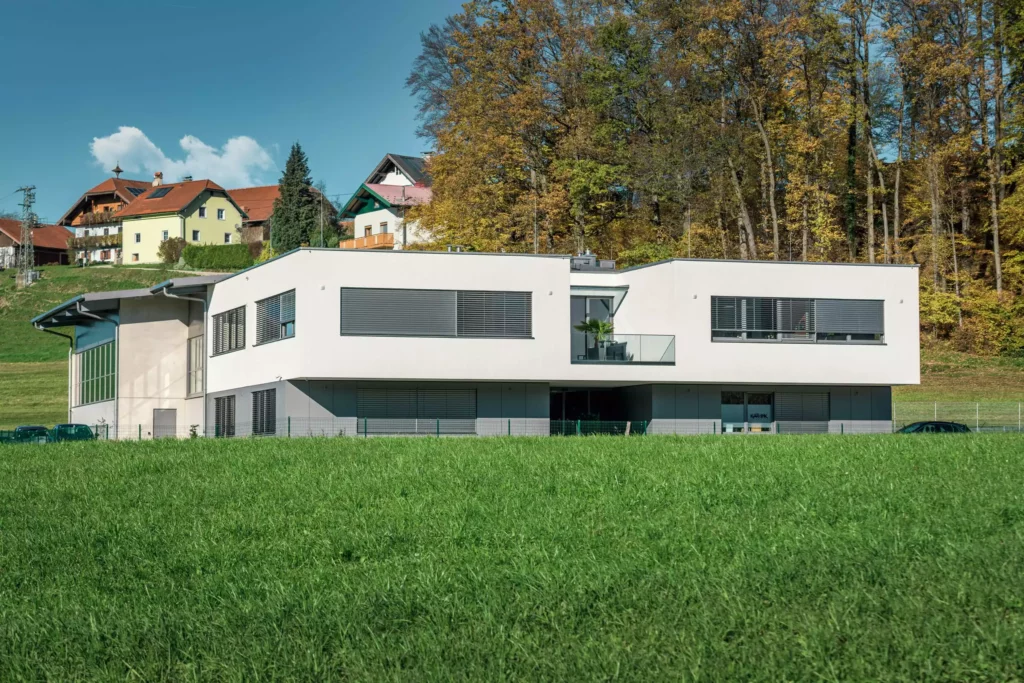 deutinger-projektentwicklung-baumanagement-generalunternehmen-immobilien-salzburg-Betriebshalle-Meister-Hallwang-9 Moderne weiße Betriebshalle mit großen Fenstern vor grünem Feld und herbstlichem Wald in Hallwang bei Salzburg